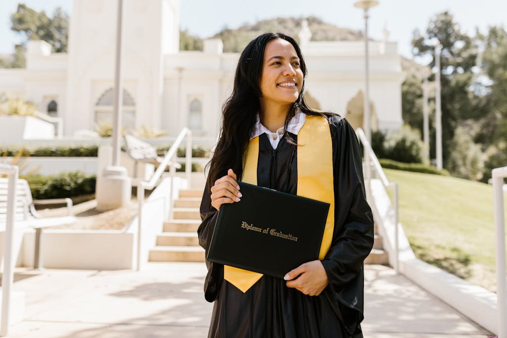 A joyful graduate in cap and gown holding a diploma on campus.