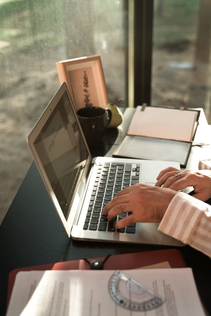 A person typing on a laptop at a sunlit desk with documents and a notebook.